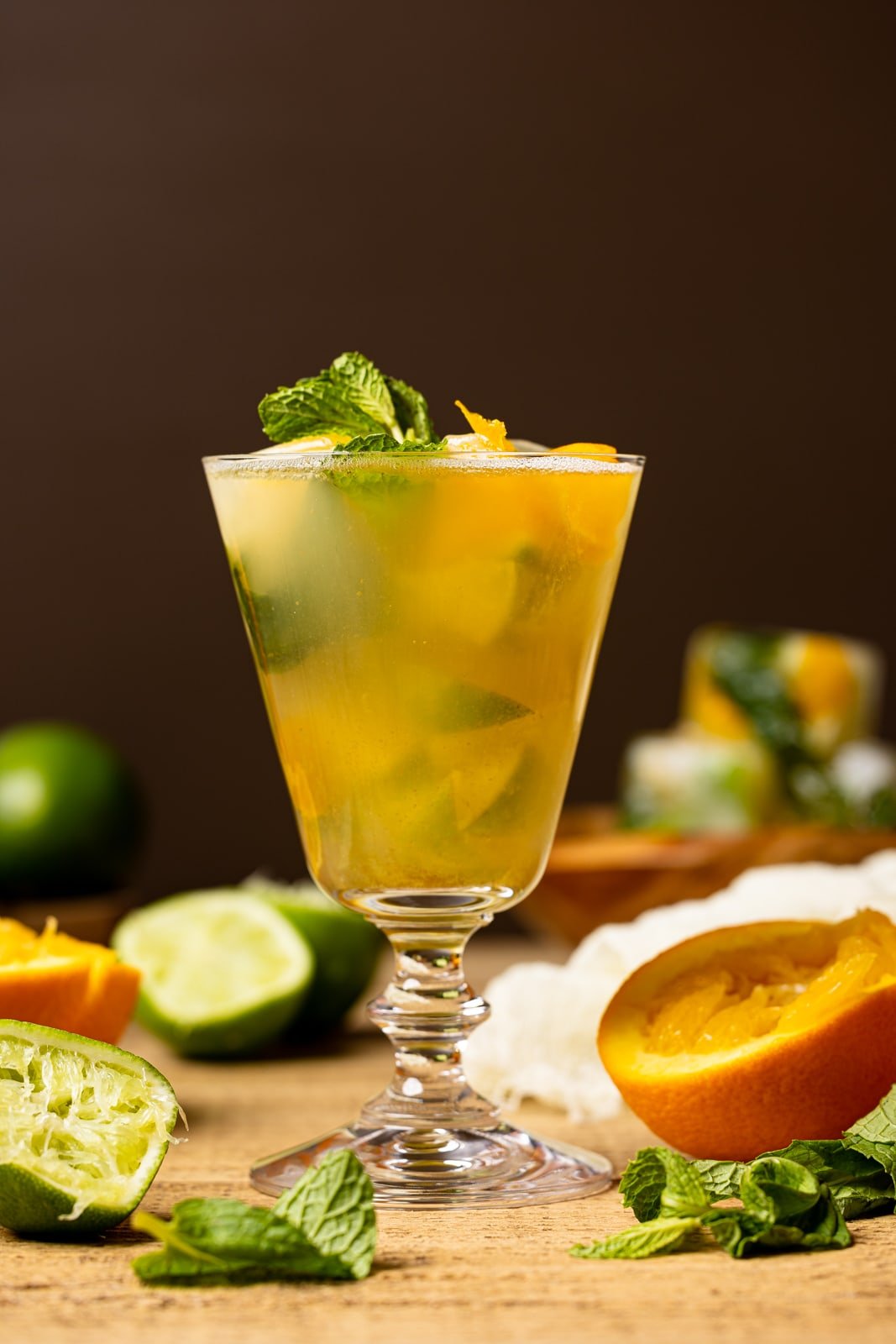 A tall glass full of mocktail mixture with slices of oranges, lime, fresh mint leaves on a brown wood table.