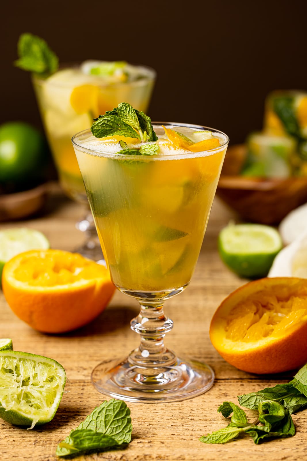 Mocktail in a tall glass on a brown wood table with halved oranges, limes, and ice cubes in the background.