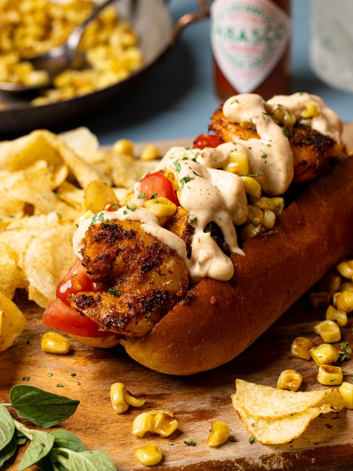 Po boy sandwich on a brown cutting board with chips and corn and hot sauce in the background.