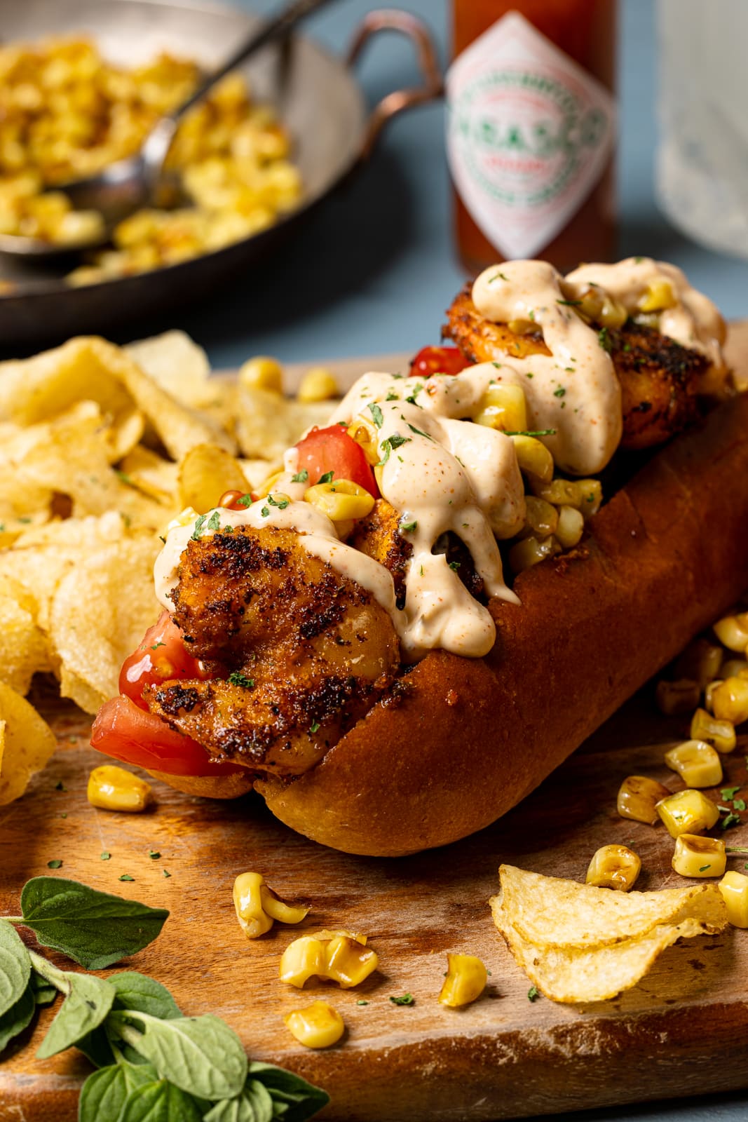 Po boy sandwich on a brown cutting board with chips and corn and hot sauce in the background.