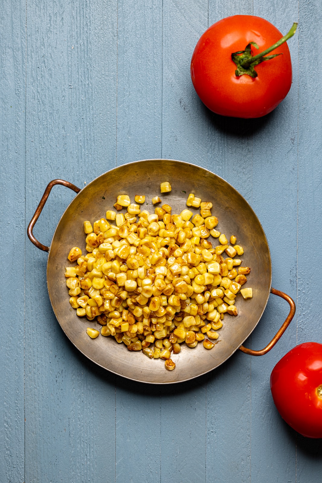 Charred corn in a bowl on a blue wood table with two tomatoes.