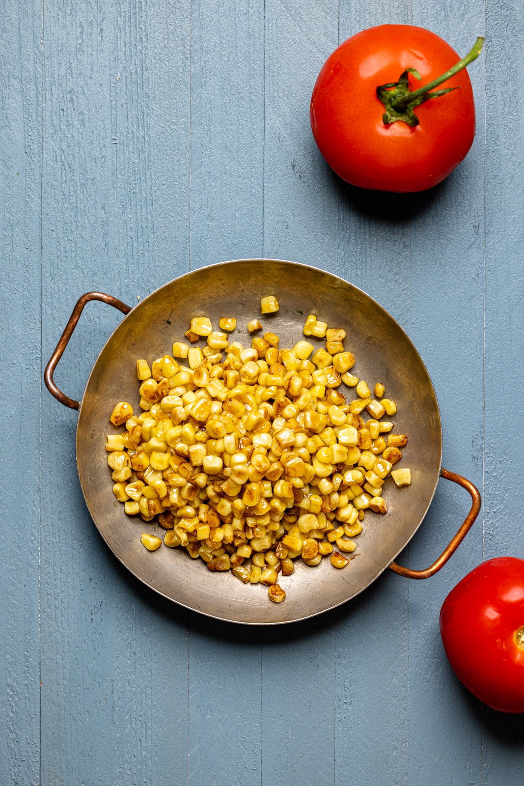 Charred corn in a bowl on a blue wood table with two tomatoes.