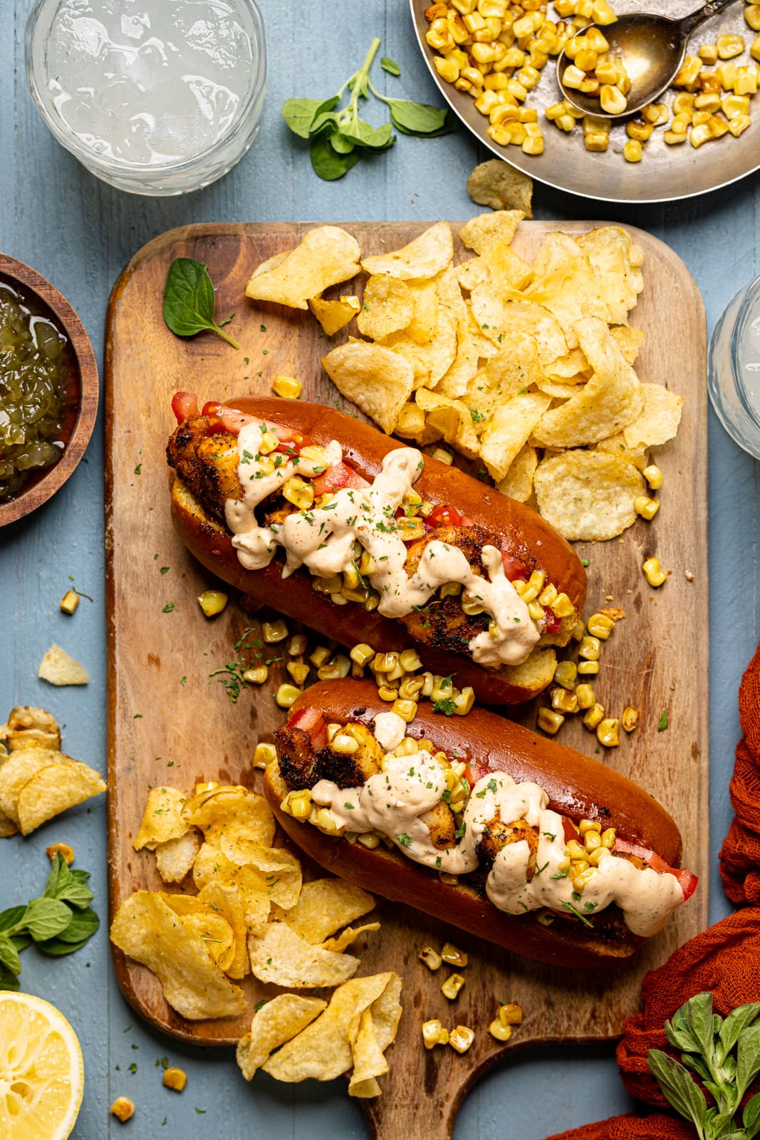 Po\' boy sandwiches on a cutting board with a side of chips, a drink, and corn on a blue wood table.