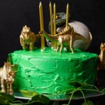 Birthday cake with green frosting and candles with toy dinosaurs on top and surrounding it on a black table.