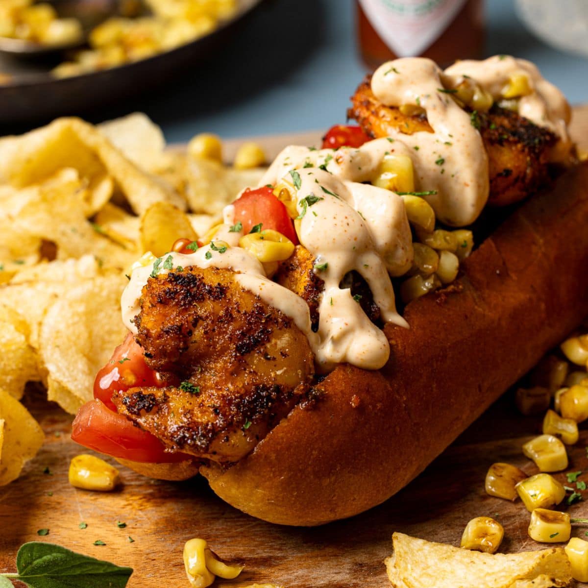 Po boy sandwich on a brown cutting board with chips and corn and hot sauce in the background.