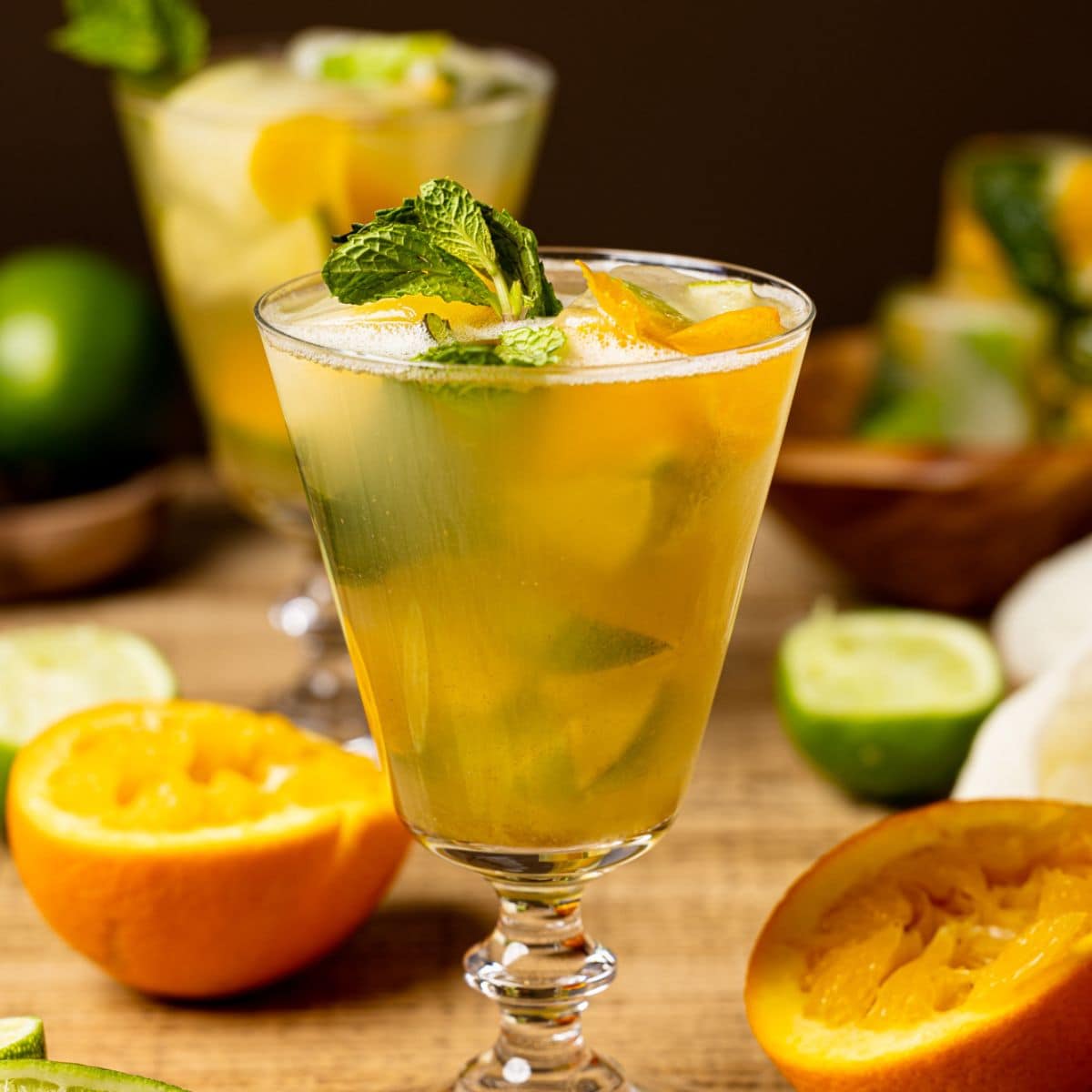 Mocktail in a two tall glasses on a brown wood table with halved oranges, limes, and ice cubes in the background.