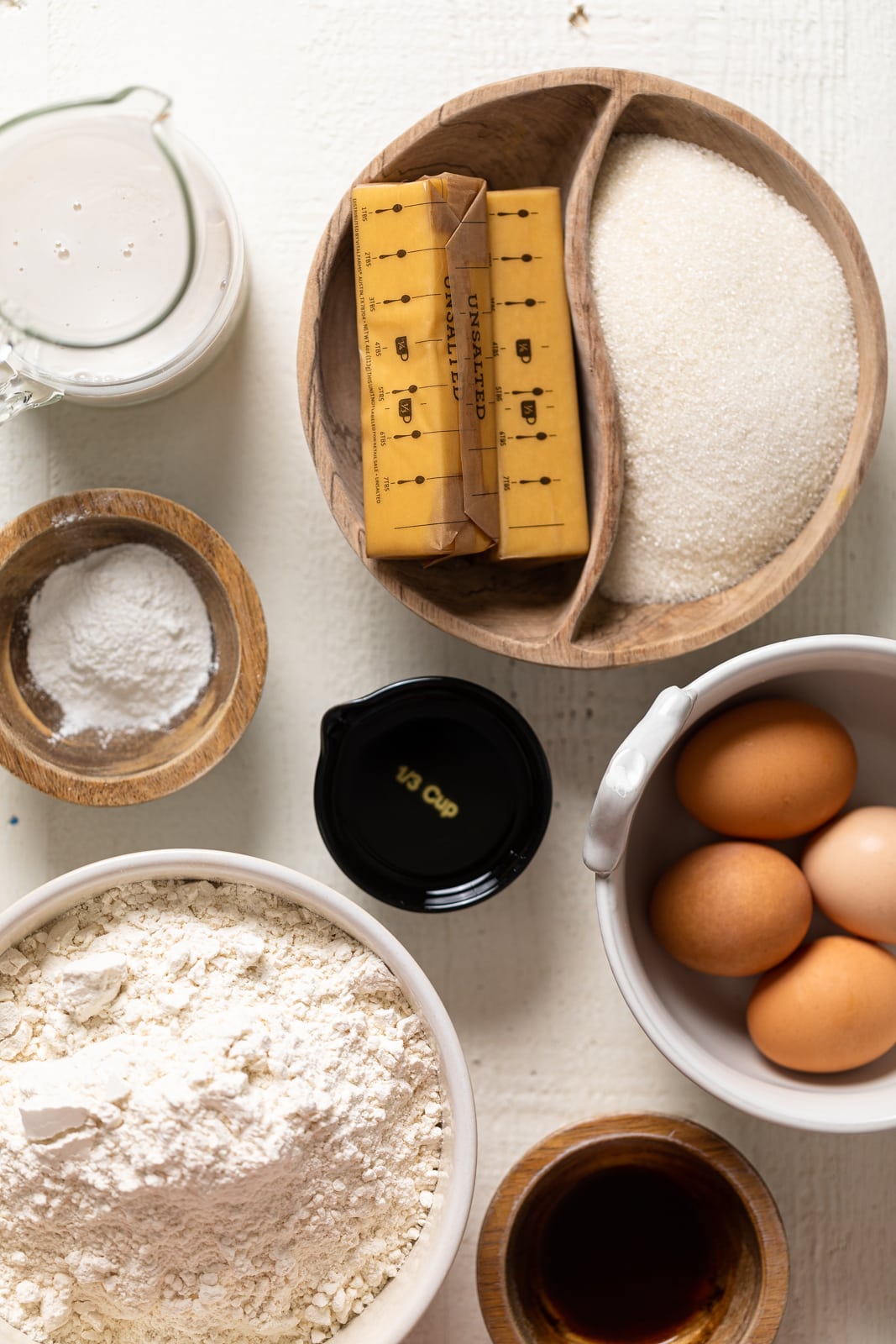 Ingredients for birthday including sugar, butter, milk, baking powder, eggs, flour, vanilla, and oil on a white wood table. 