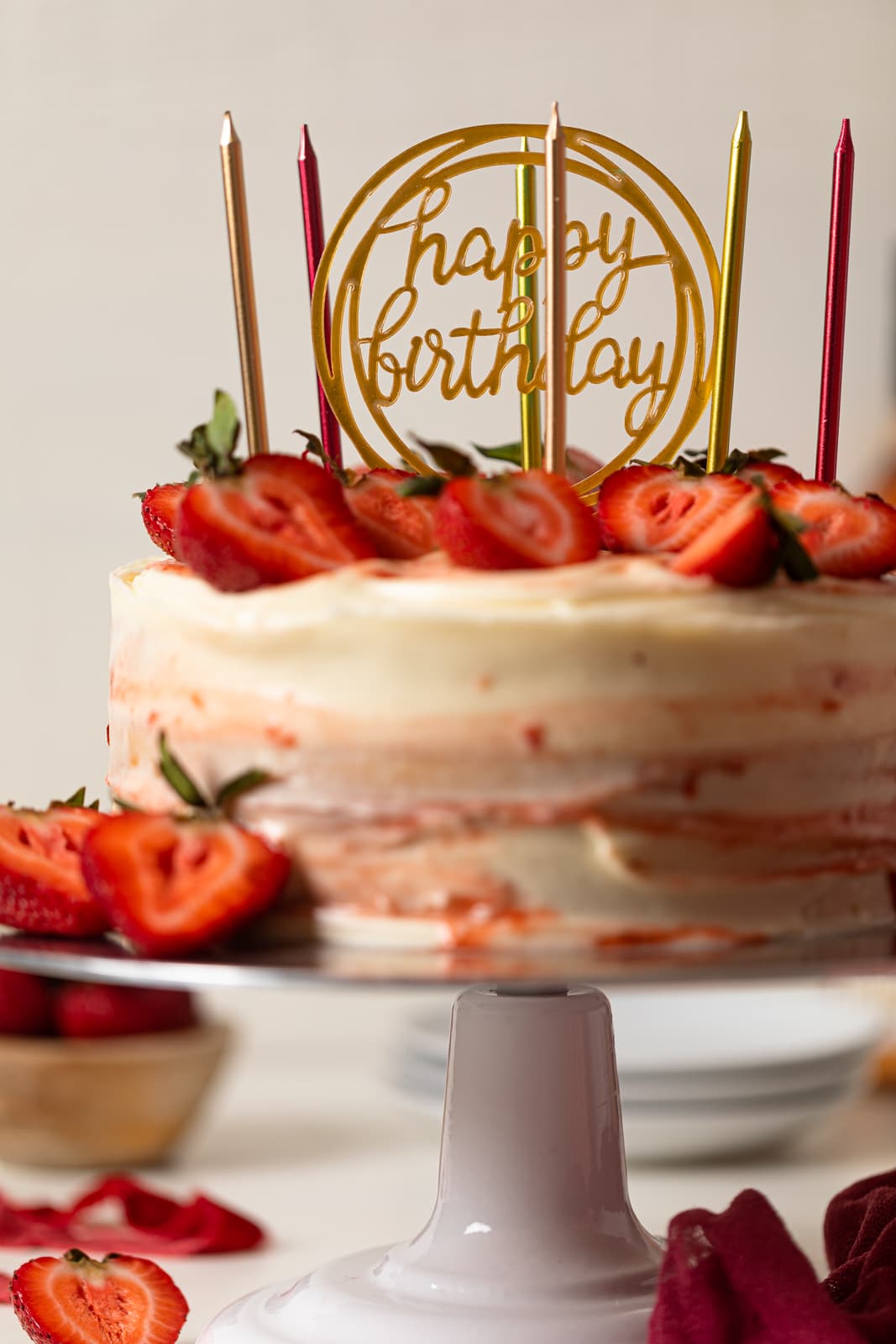 Strawberry shortcake birthday cake with sliced strawberries on top, birthday candles, on a cake stand on a white table.