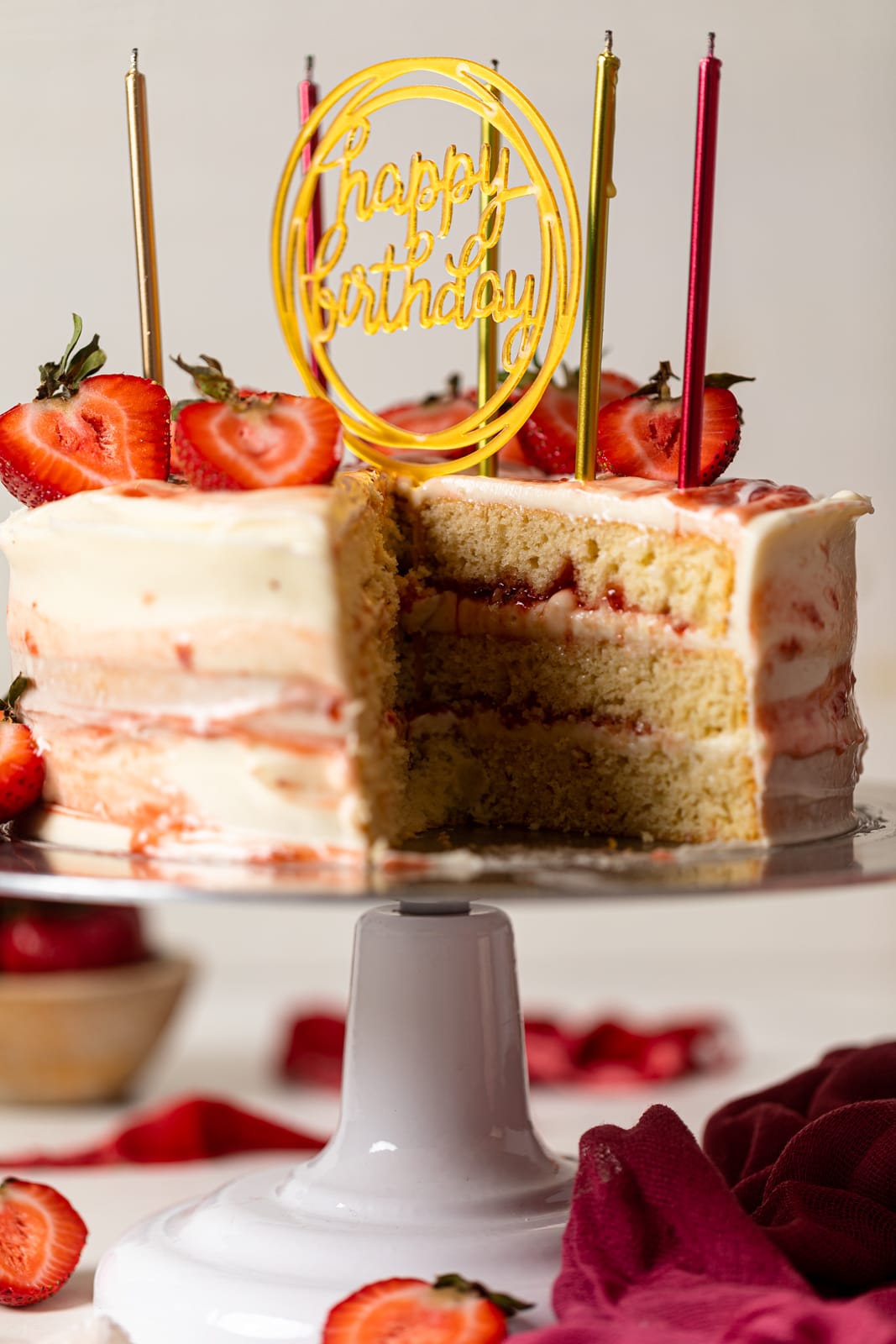 Strawberry shortcake birthday cake with a slice removed and sliced strawberries on top, birthday candles, on a cake stand on a white table.