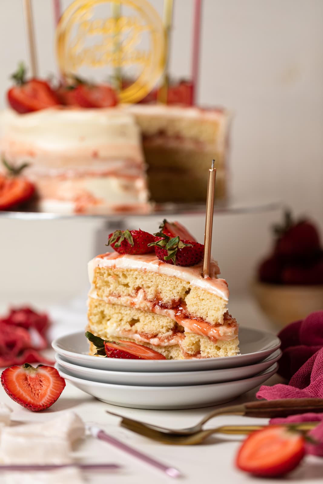 Slice of cake on stacked white plates with sliced strawberries and the full cake on a cake stand in the background; all on a white table. 