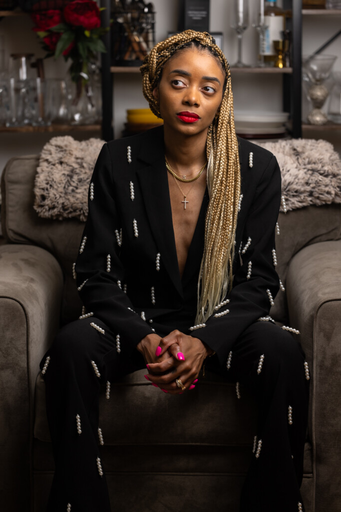 Shanika sitting upward on a couch in a black + white suit with hands in front and looking slightly to side.