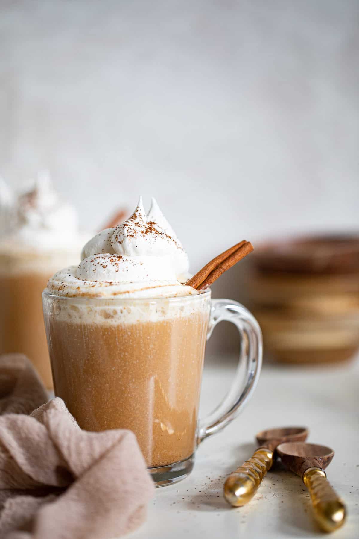 Glass mug of Warm Apple Chai Latte topped with whipped cream and a cinnamon stick.