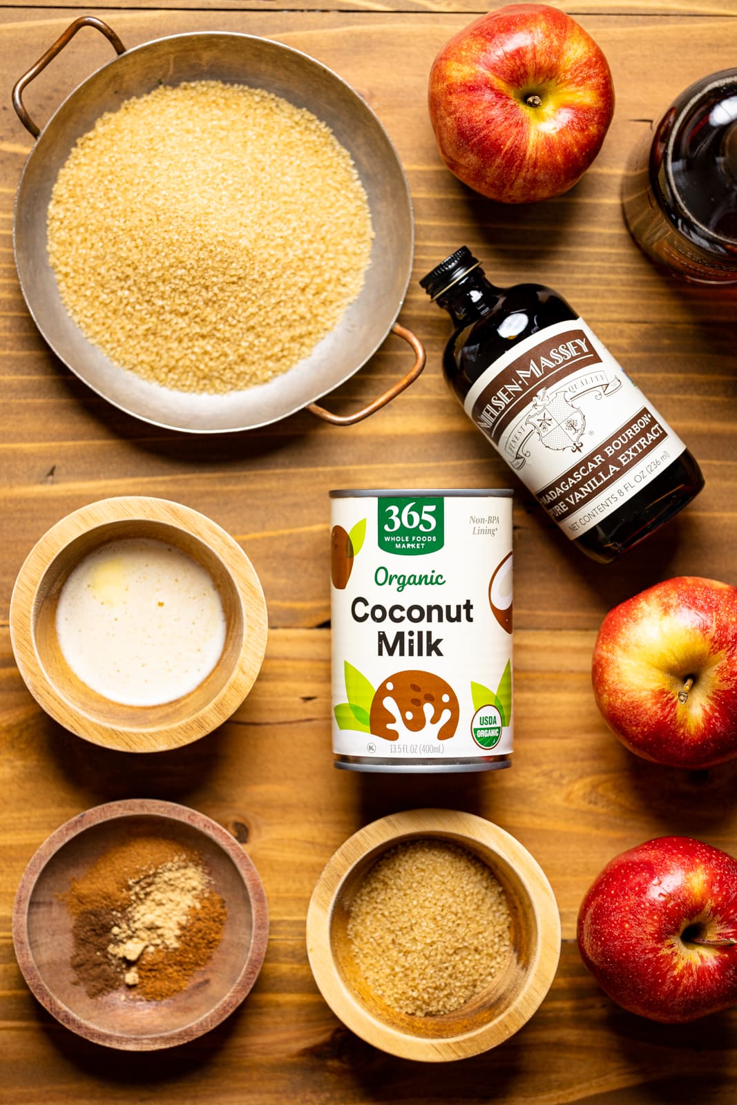 Ingredients on a brown wood table including sugar, apples, coconut milk, spices, vanilla, maple syrup, and melted butter.