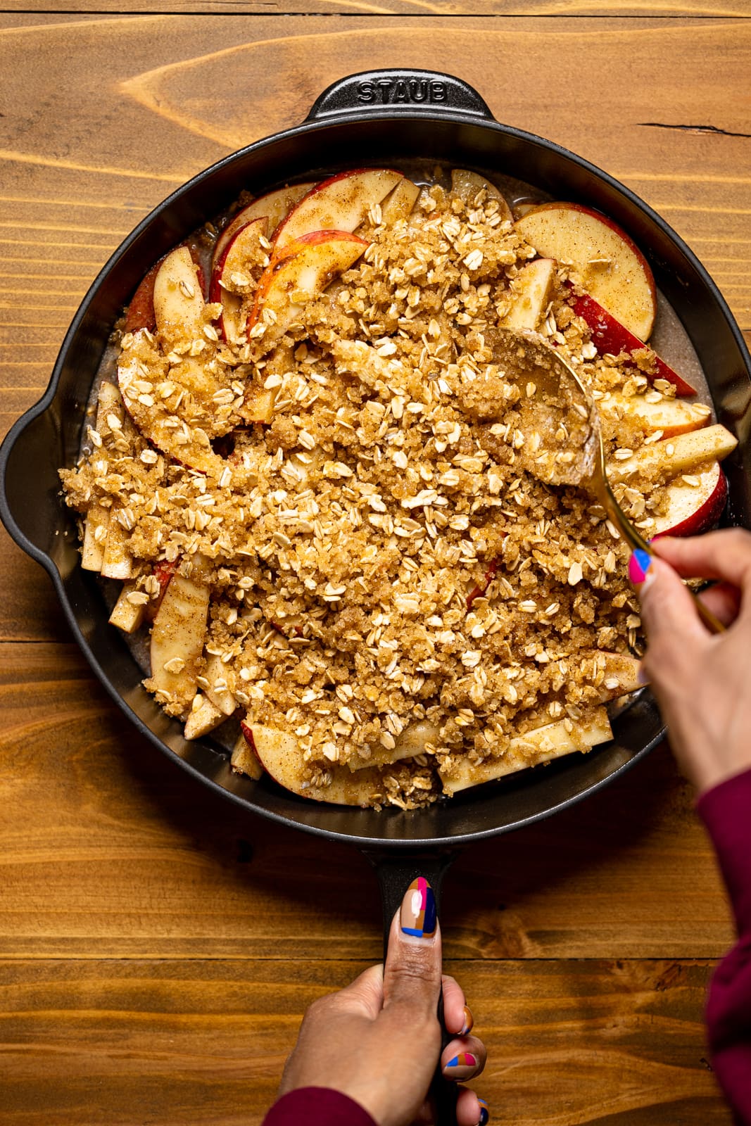 Crisp topping being spooned unto apple mixture in a black skillet on a brown wood table.