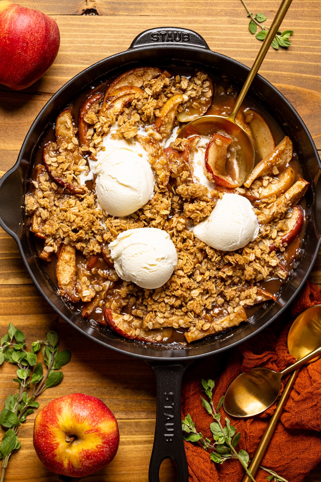 Baked apple crisp in a black skillet with scoops of ice cream, spoons, apples, and herbs.