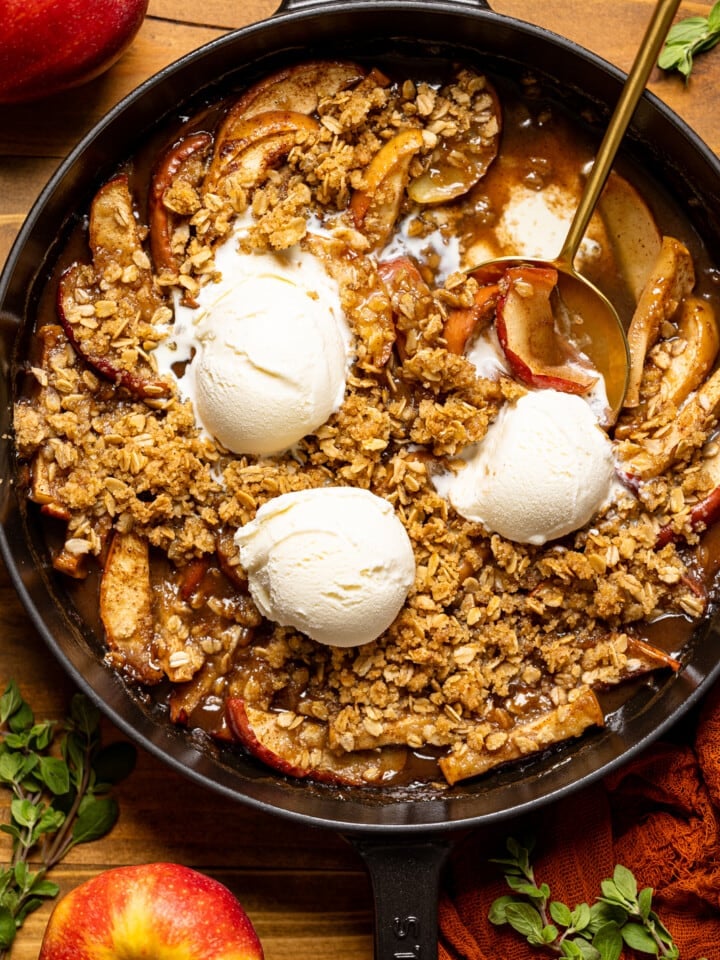 Baked apple crisp in a black skillet with scoops of ice cream, spoons, apples, and herbs.