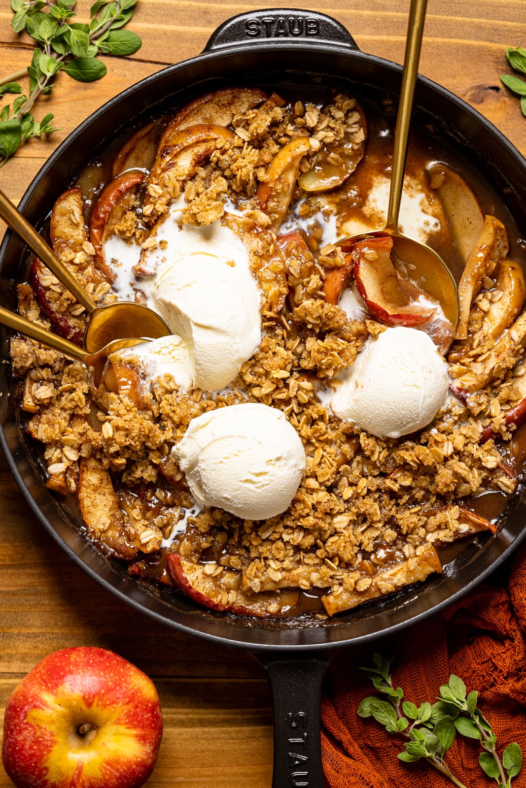 Baked apple crisp in a black skillet with scoops of ice cream, spoons, apples, and herbs.