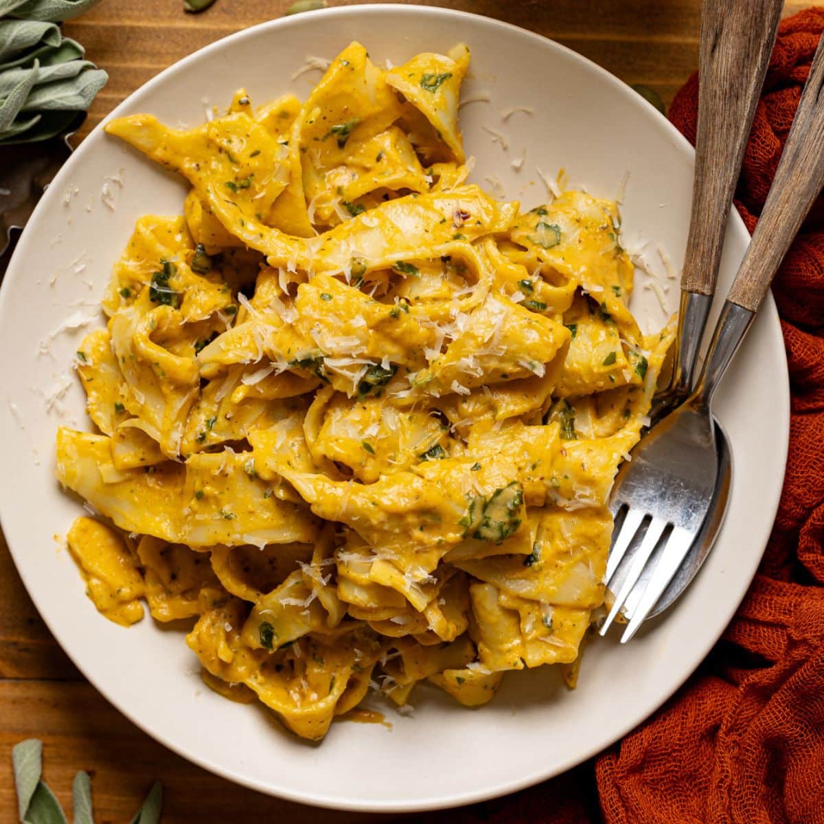 Creamy pasta on a white plate with a fork and spoon along with a glass of juice and pumpkin seeds.