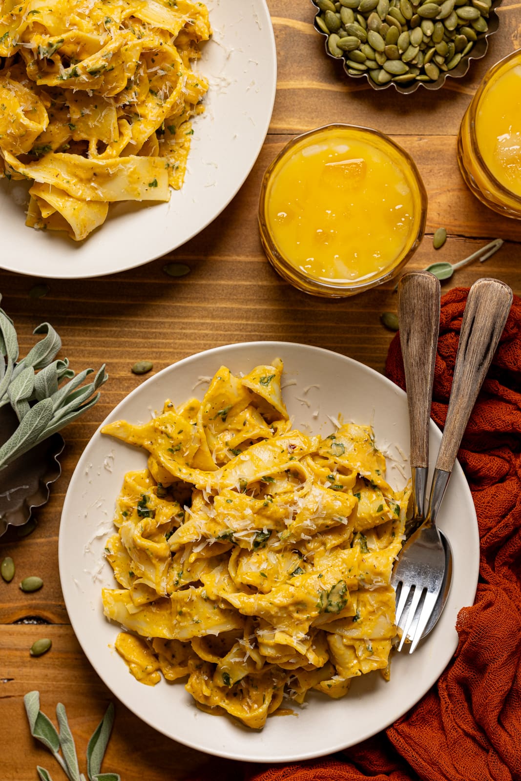 Two plates of pasta with a fork, spoon, and glasses of juice with pumpkin seeds.