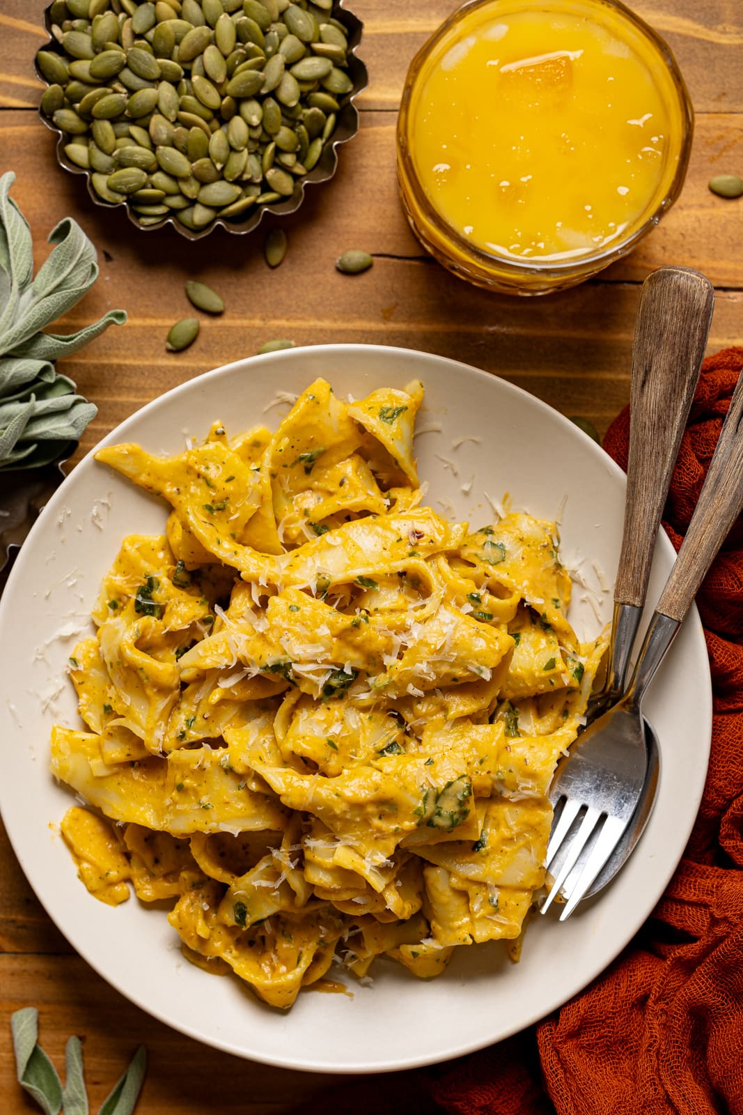 Creamy pasta on a white plate with a fork and spoon along with a glass of juice and pumpkin seeds.