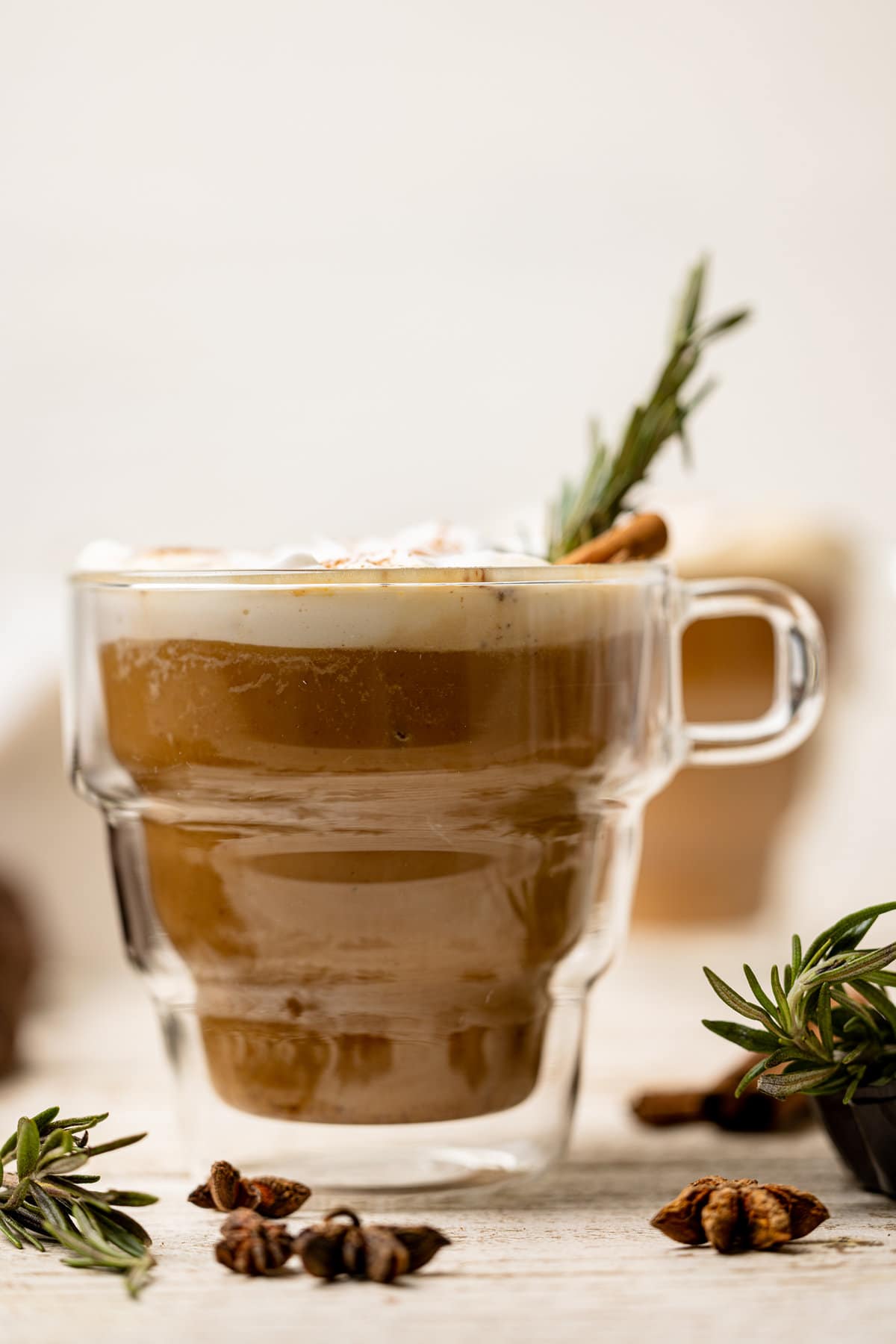 Cinnamon stick and sprig of rosemary in a glass mug of Vegan Pumpkin Chai Latte.