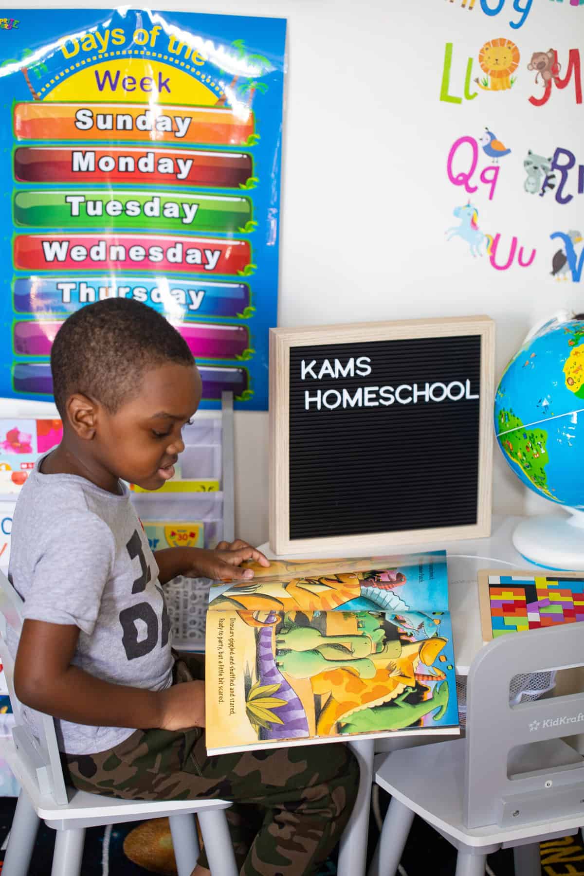 Boy reading a dinosaur book at a table with a sign that says \"Kams Homeschool.\"