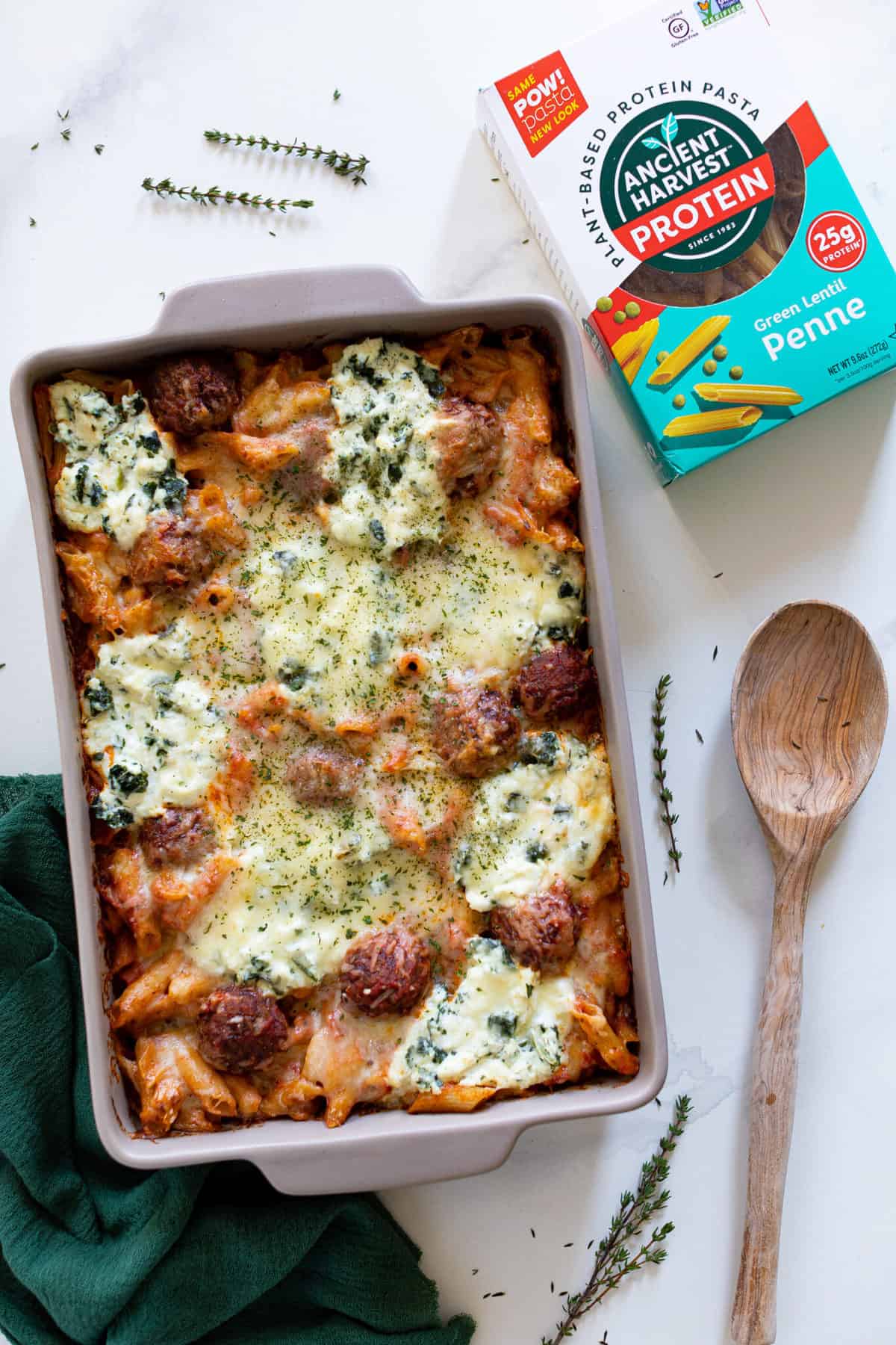 Baking dish of gluten-free Baked Meatball Penne Pasta Casserole with a wooden spoon and box of Ancient Harvest pasta.