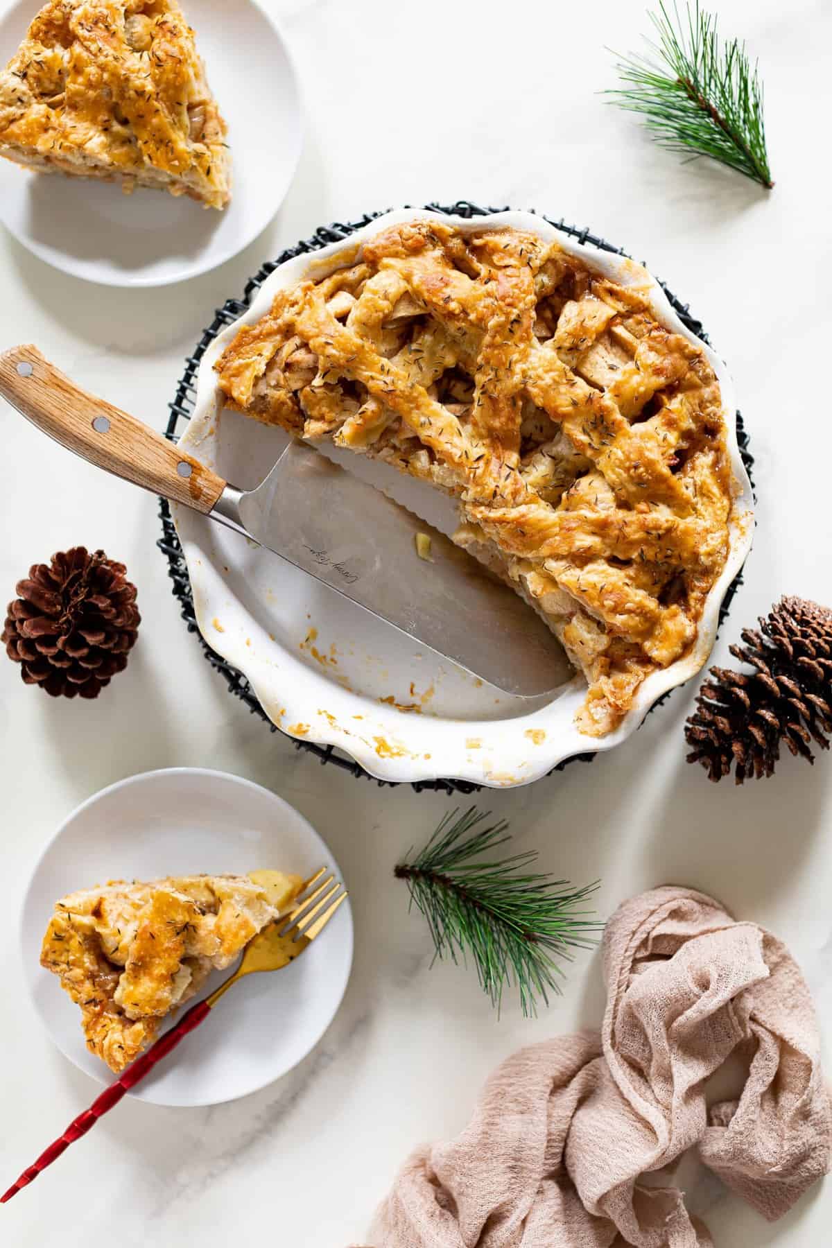 Half-served Apple Cheddar Thyme Pie on a table with pinecones and evergreen sprigs.