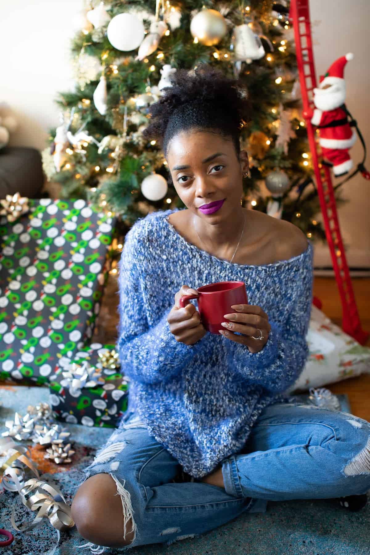 Shanika sitting in front of a Christmas tree, wearing a blue sweater, and holding a red mug.