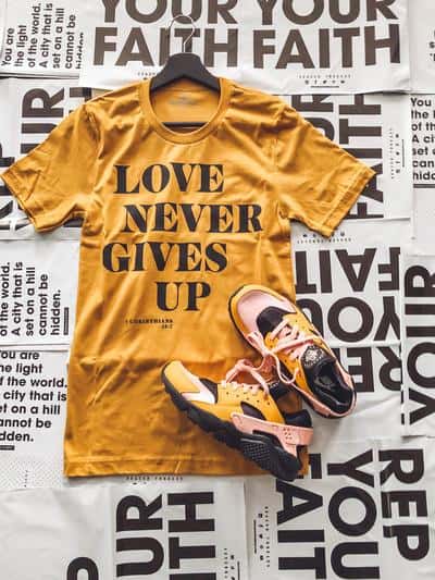 Mustard-colored shirt that reads \"Love Never Gives Up\" along with a pair of shoes.