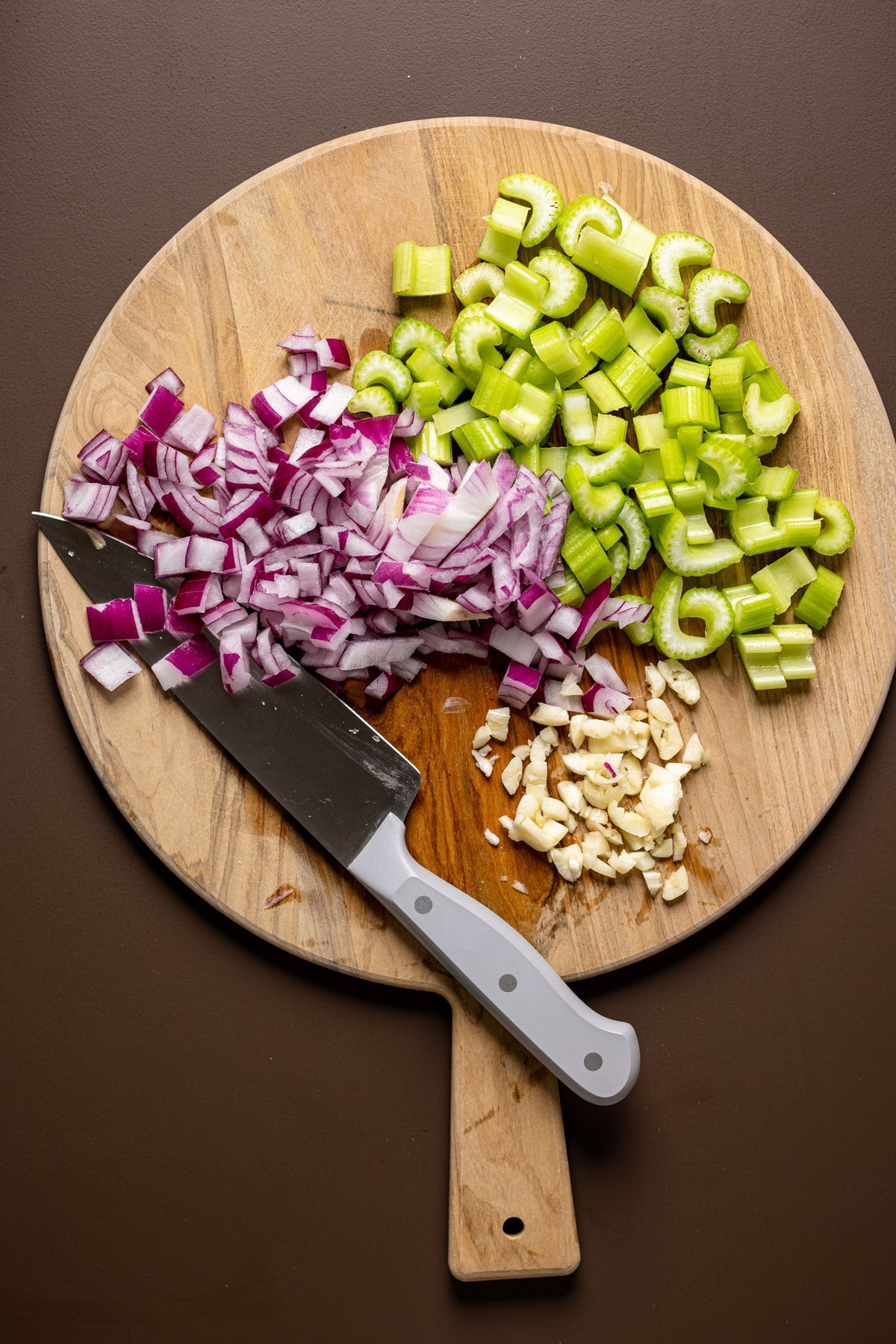 Chopped garlic, red onion, and celery on a cutting board with a knife