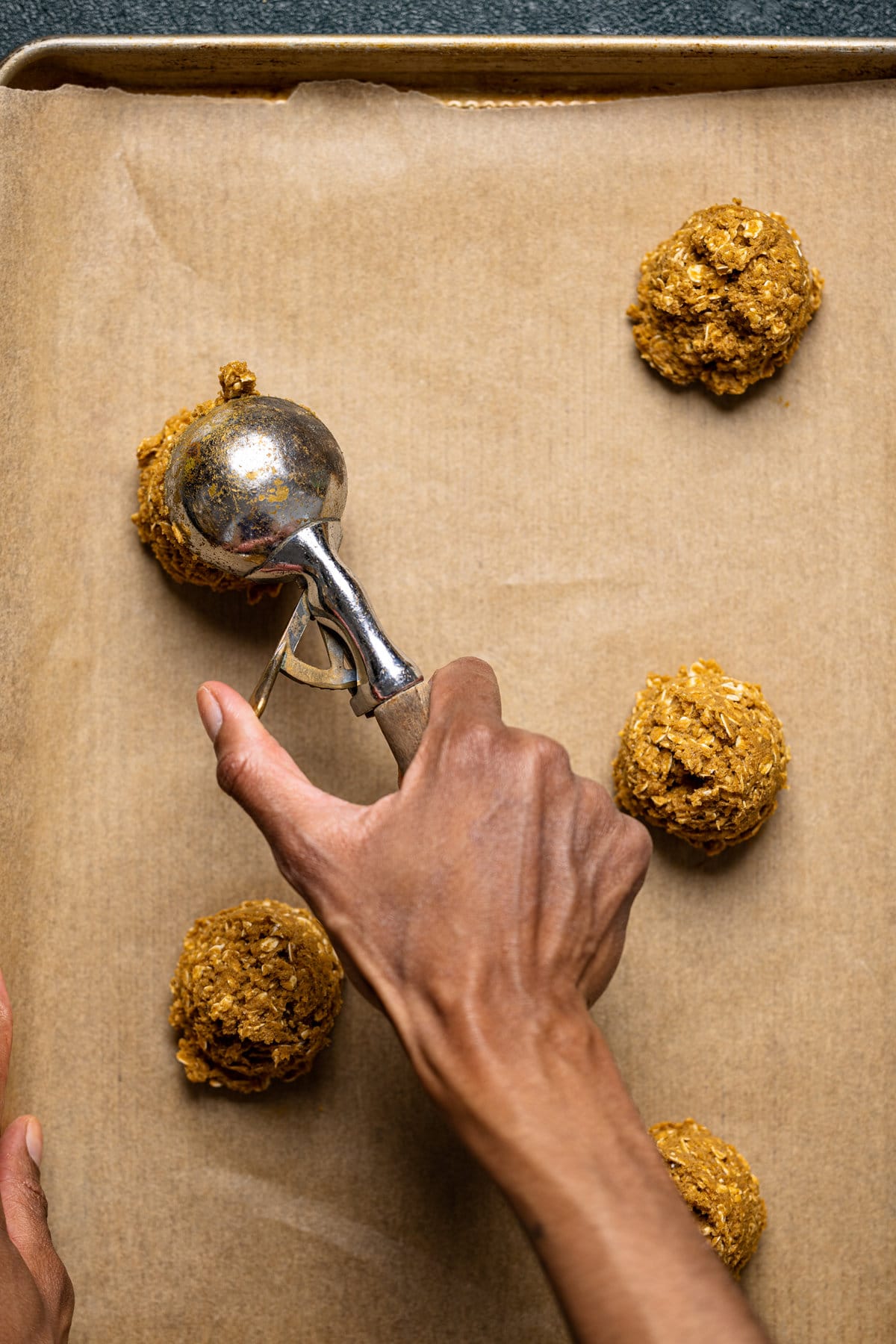 Hand using a scooper to create balls of Gingerbread Oatmeal Cookie dough on parchment paper