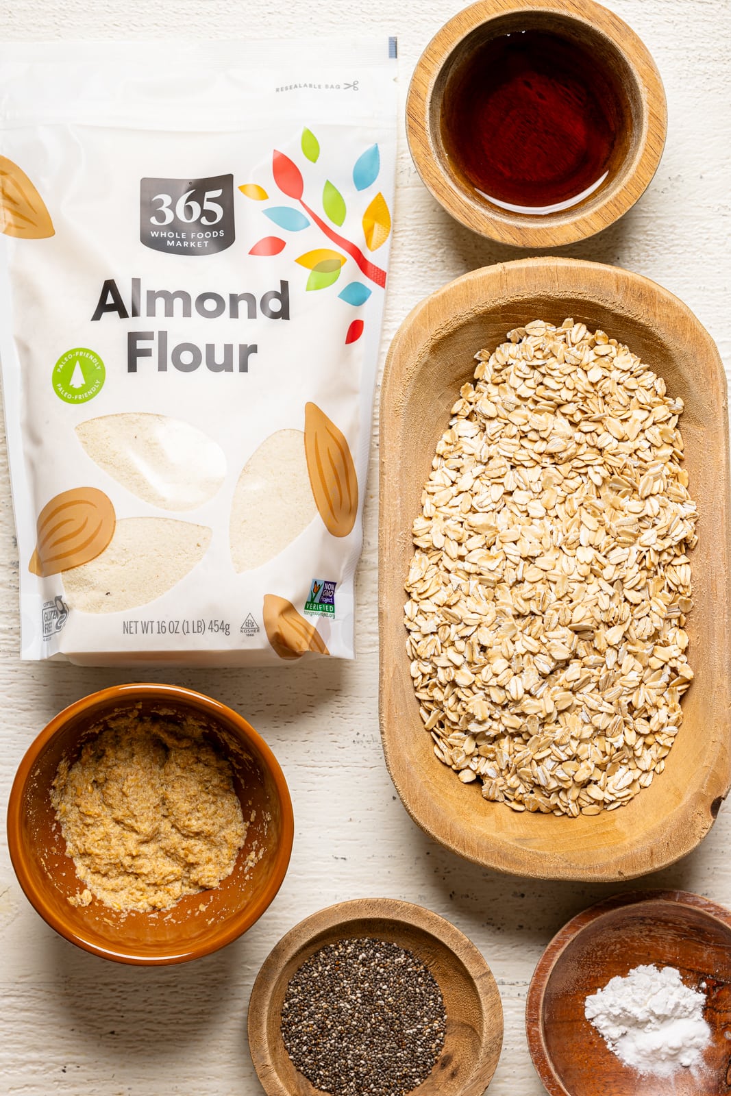 Ingredients on a white table including flour, oats, flax eggs, maple syrup, chia seeds, and baking powder. 