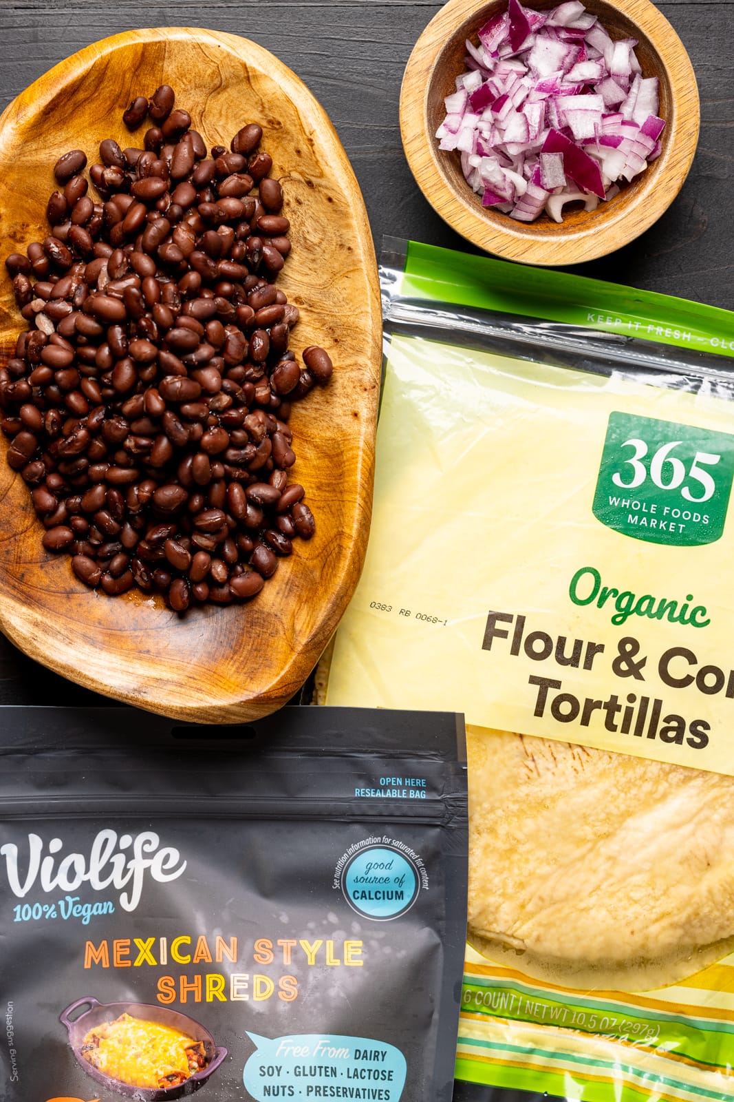 Ingredients on a black wood table including tortillas, black beans, cheese, and onions.