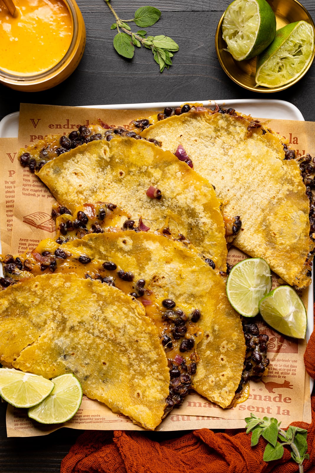 Baked tacos on a white plate with chipotle sauce, lime wedges, red napkin, and herbs.