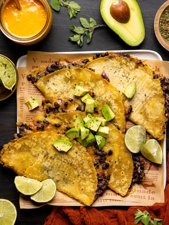 Baked tacos on a white plate with lime wedges, chipotle sauce, avocado, red napkin, and herbs.