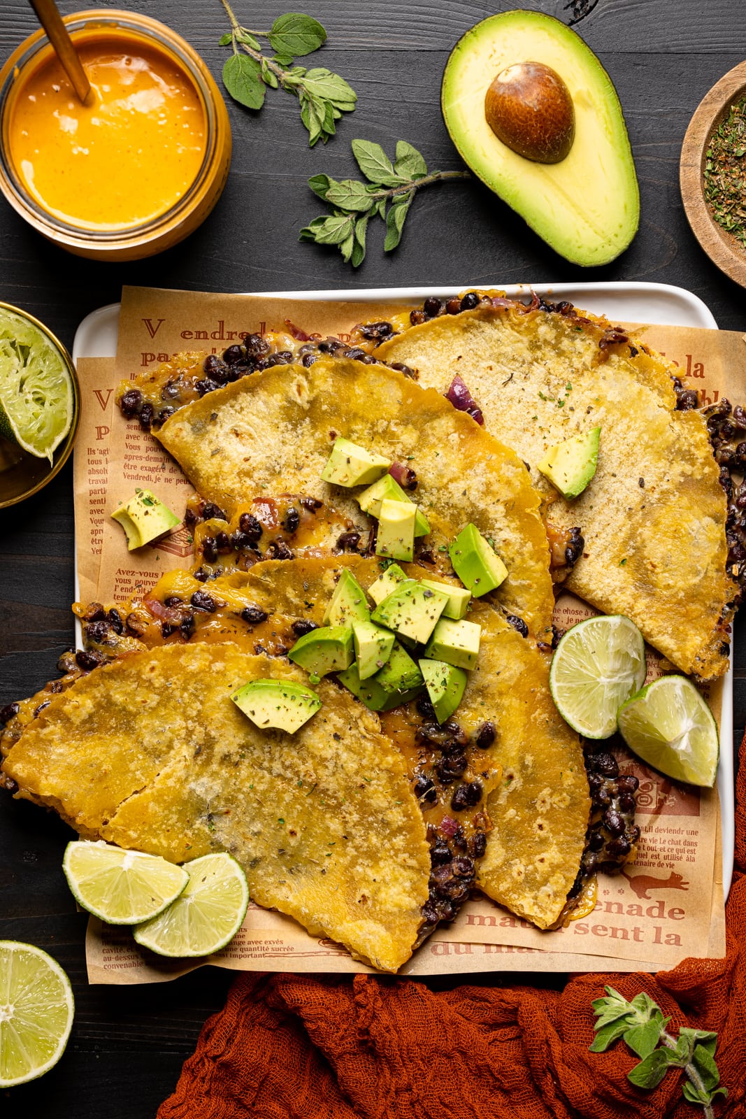 Baked tacos on a white plate with lime wedges, chipotle sauce, avocado, red napkin, and herbs.