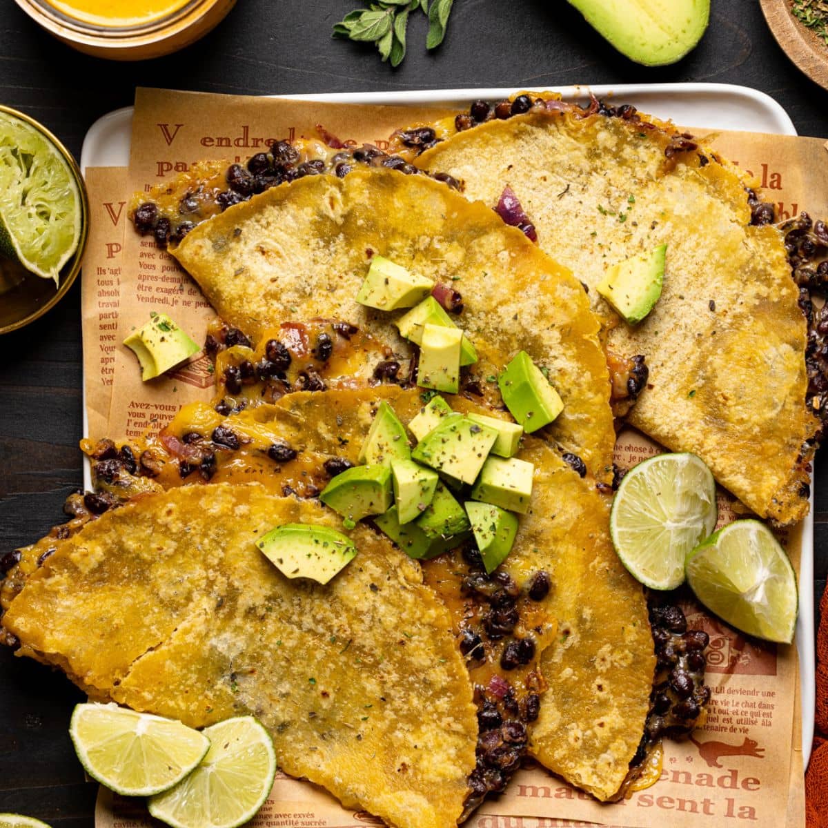 Baked tacos on a white plate with lime wedges, chipotle sauce, avocado, red napkin, and herbs.