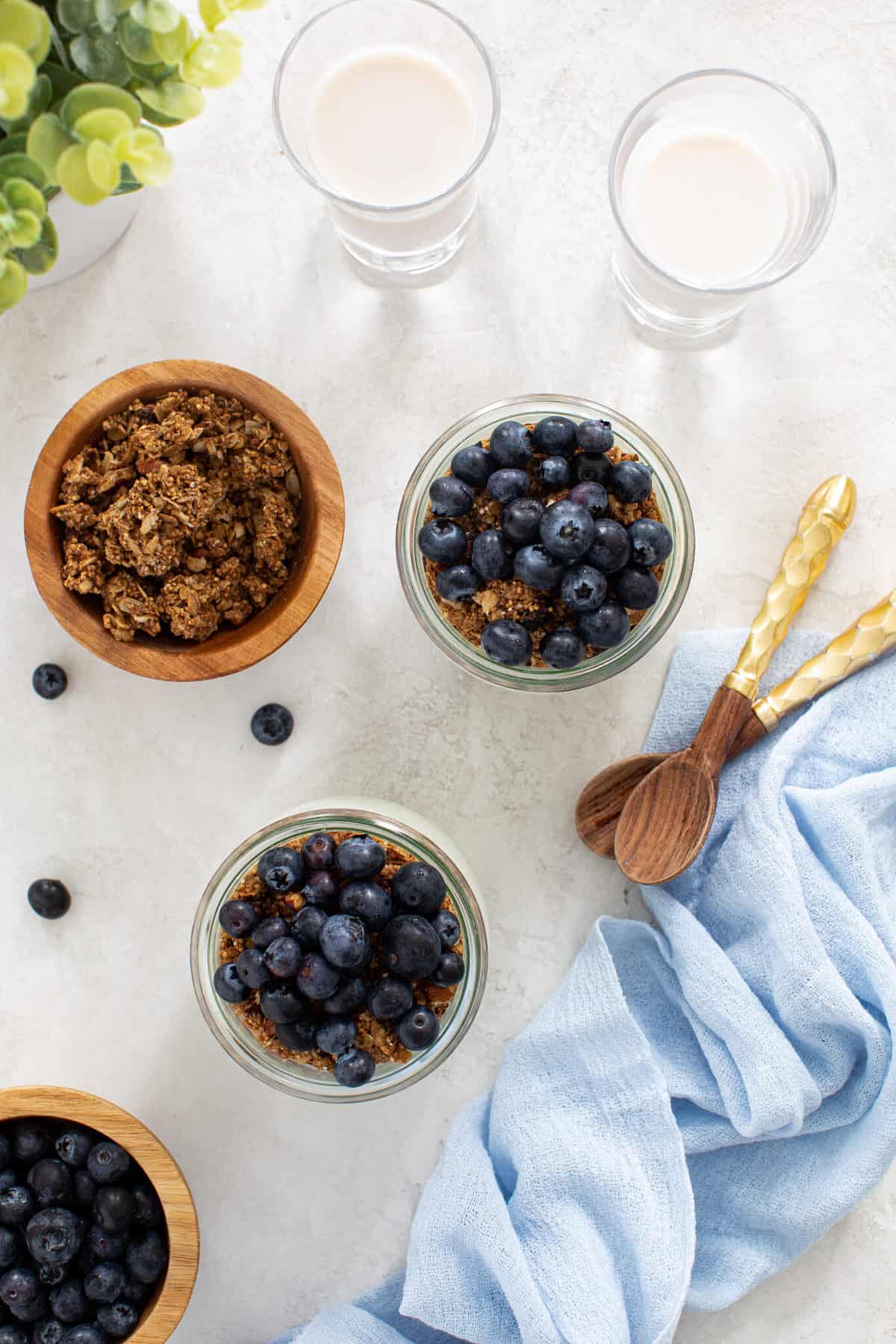 Overhead of two blueberry parfaits with dairy free yogurt and granola with two glasses of milk and wooden spoons.