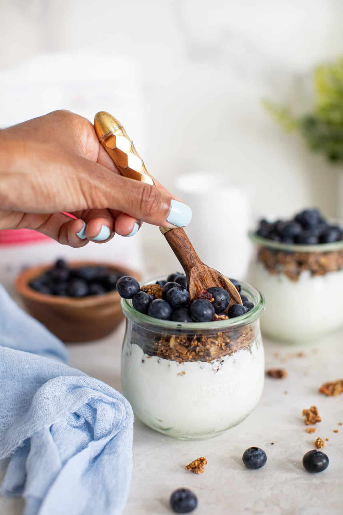 Close up on a spoon taking a scoop out of blueberry breakfast parfait with granola in a clear glass jar.