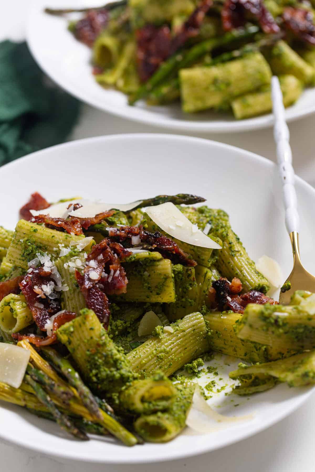 Pesto Pasta with Asparagus + Bacon on a white plate with a gold and white fork taking a bite