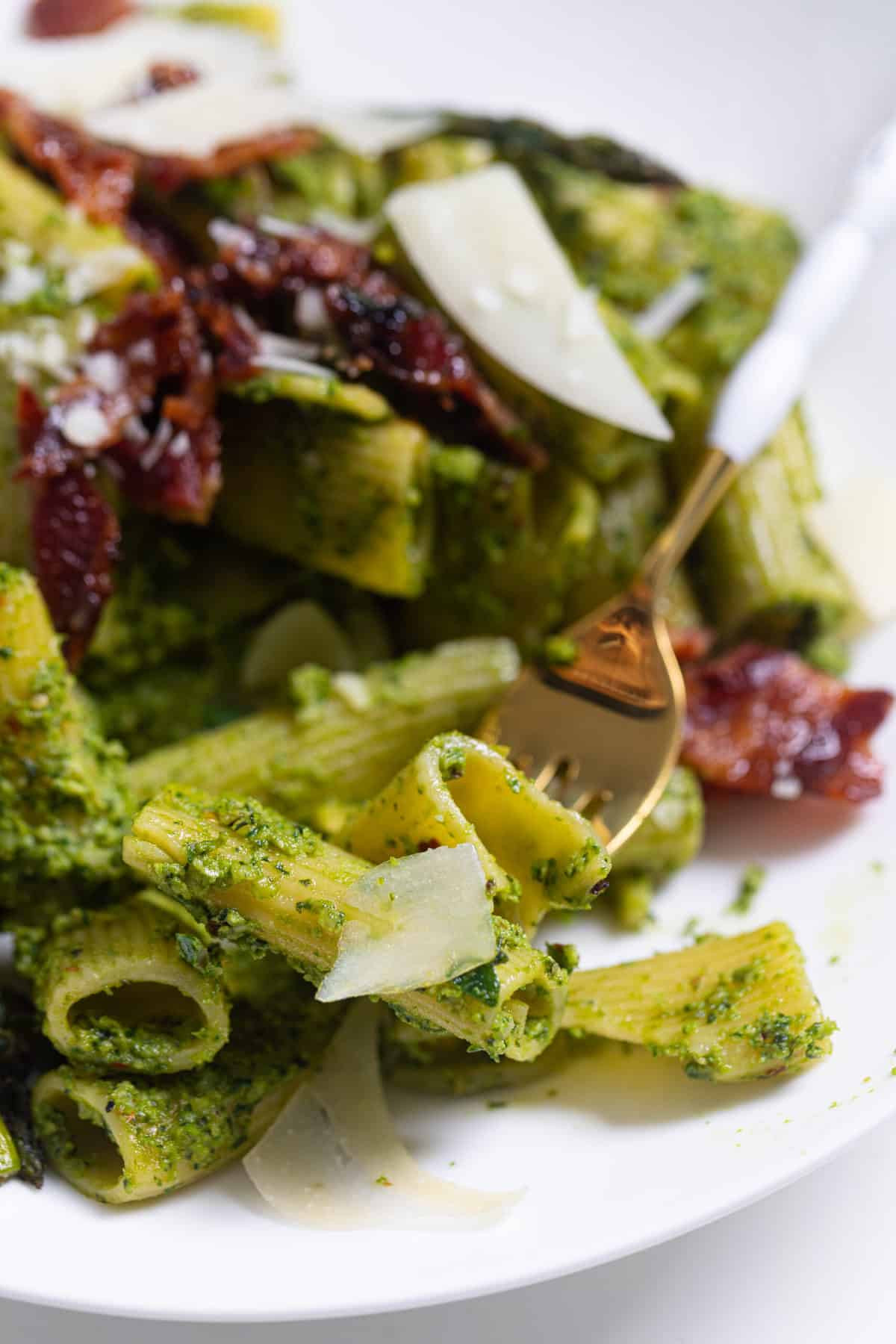 Pesto Pasta with Asparagus + Bacon on a white plate with a gold and white fork takign a bite