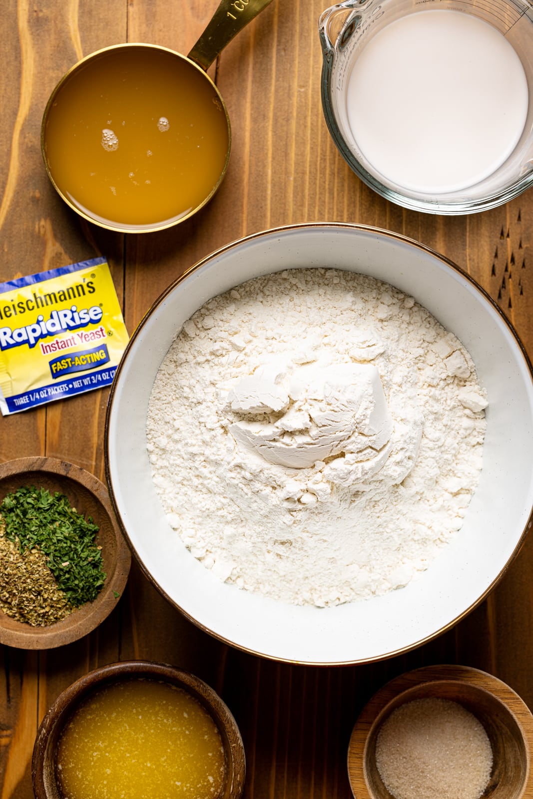 Ingredients on a brown wood table including flour, yeast, seasonings and herbs, milk, melted butter, and sugar.