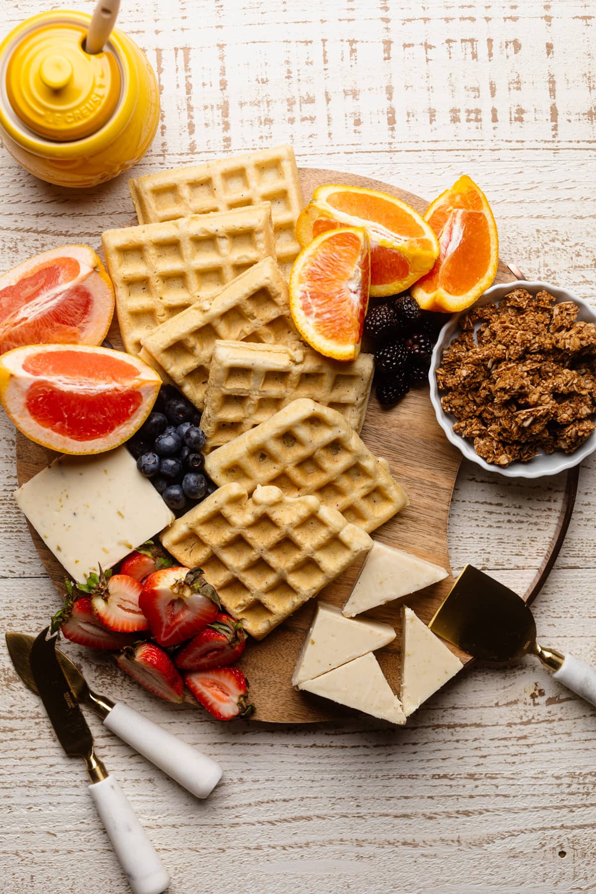 Fruits, waffles, vegan cheese, and a bowl of granola on a wooden board.