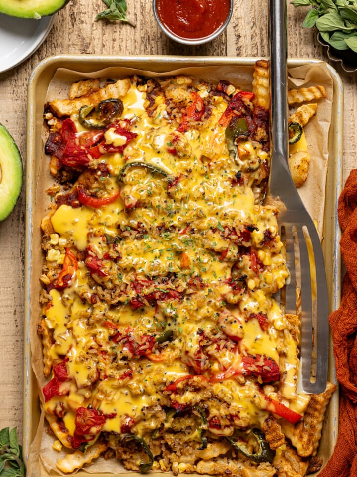 Loaded fries in a sheet pan with a spatula, avocado, and a side of ketchup on a wood table.