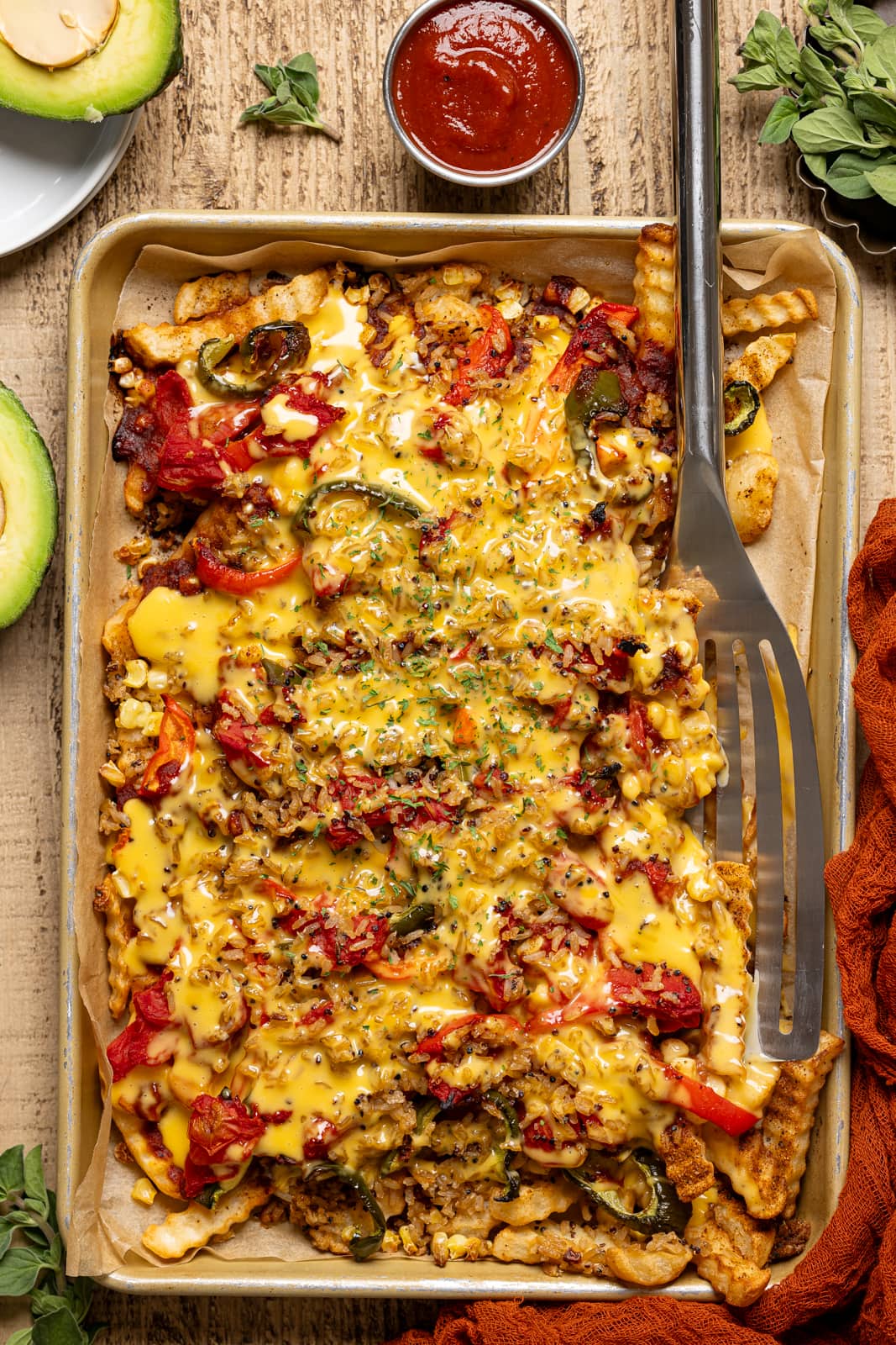 Loaded fries in a sheet pan with a spatula, avocado, and a side of ketchup on a wood table.