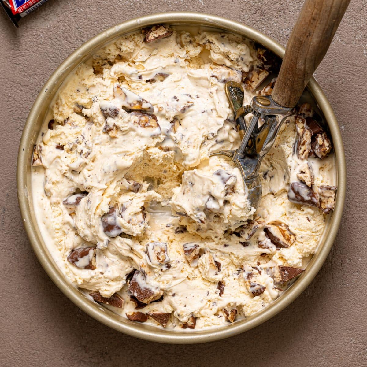 Snickers ice cream in a round metal pan on a greyish brown table with a scoop and candy bar.