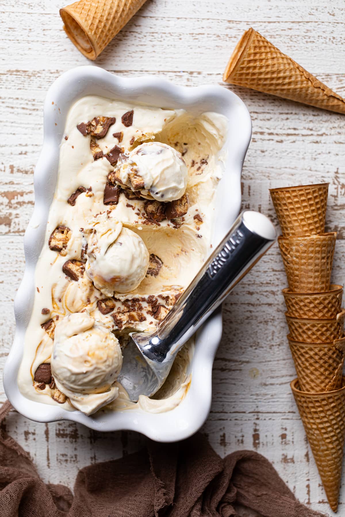 Stack of ice cream cones next to a container of Loaded No-Churn Snickers Ice Cream.