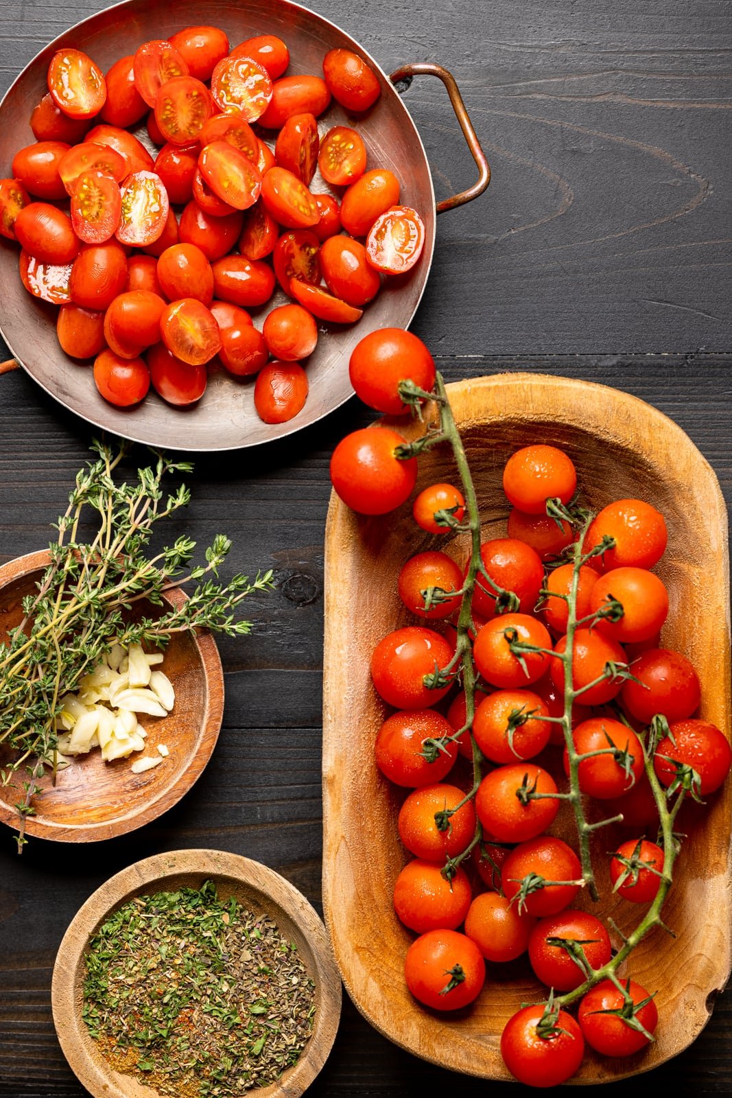 Two kinds of tomatoes in bowls on a black wood table along with herbs + seasonings.