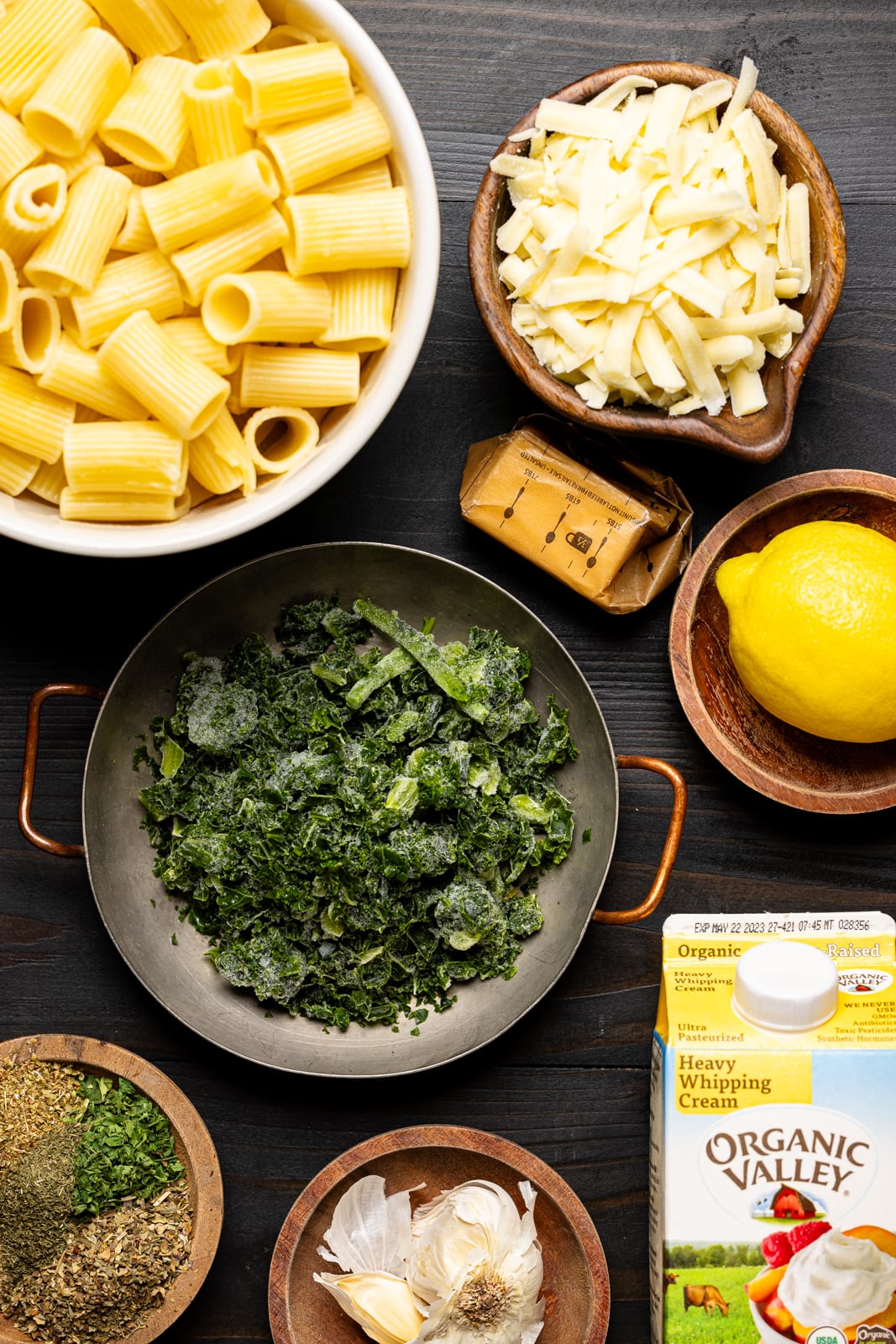 Ingredients on a black table including rigatoni pasta, kale, heavy cream, herbs + seasonings, butter, lemon, garlic, and cheese.